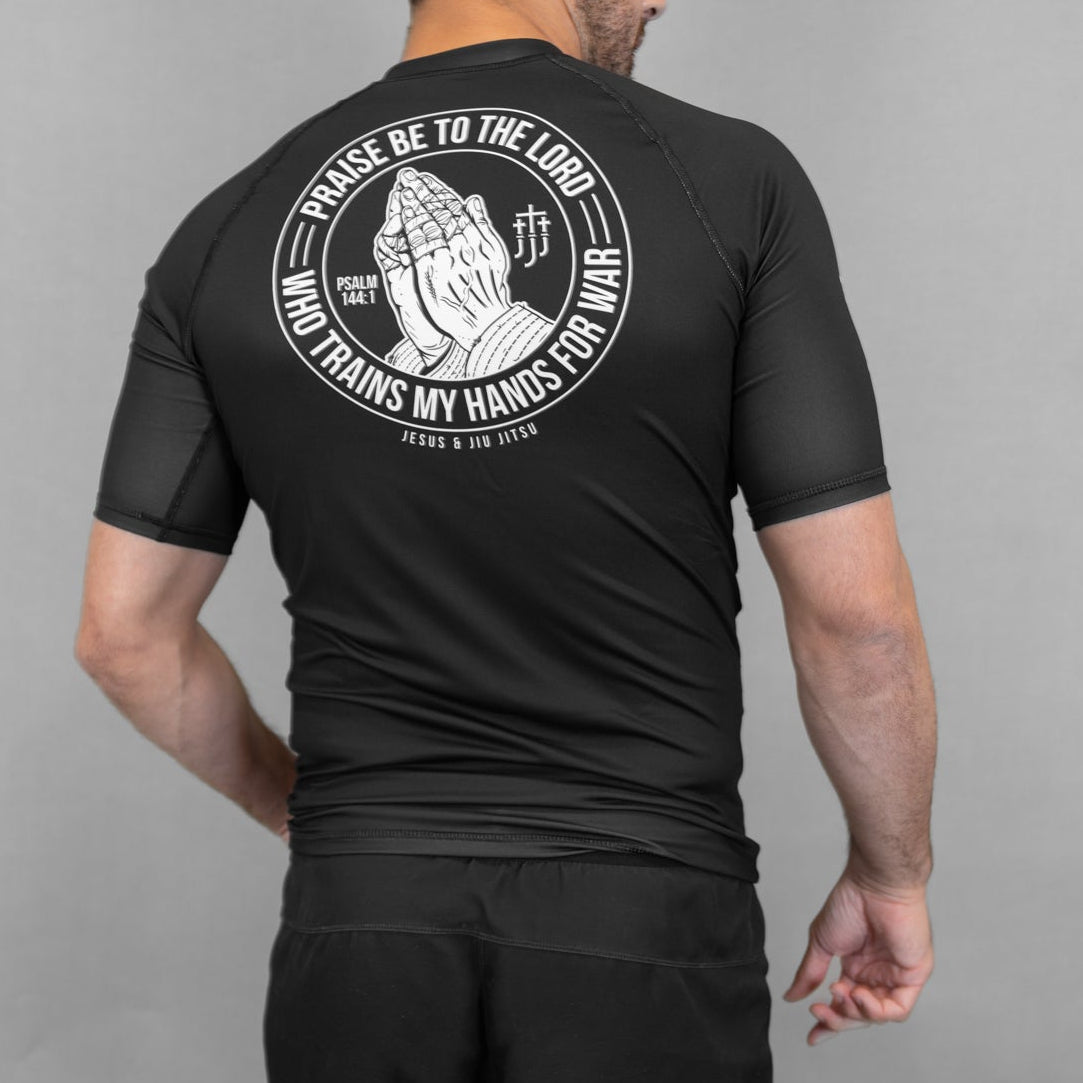 Person wearing a black Prayer hands rashguard and shorts with a logo on the back against a gray background