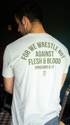 A man viewed from behind wearing a sand t-shirt with olive green text on the back that reads 'FOR WE WRESTLE NOT AGAINST FLESH & BLOOD EPHESIANS 6:12' and a small monogram logo below.